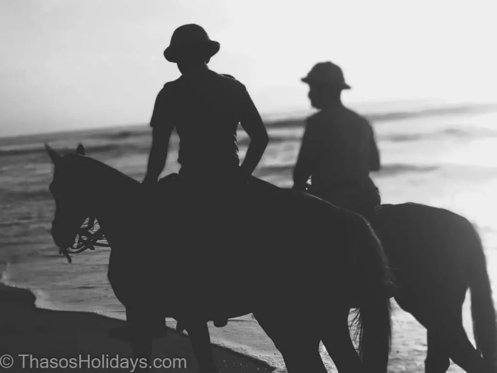 Two horses with people on top walking near the beach